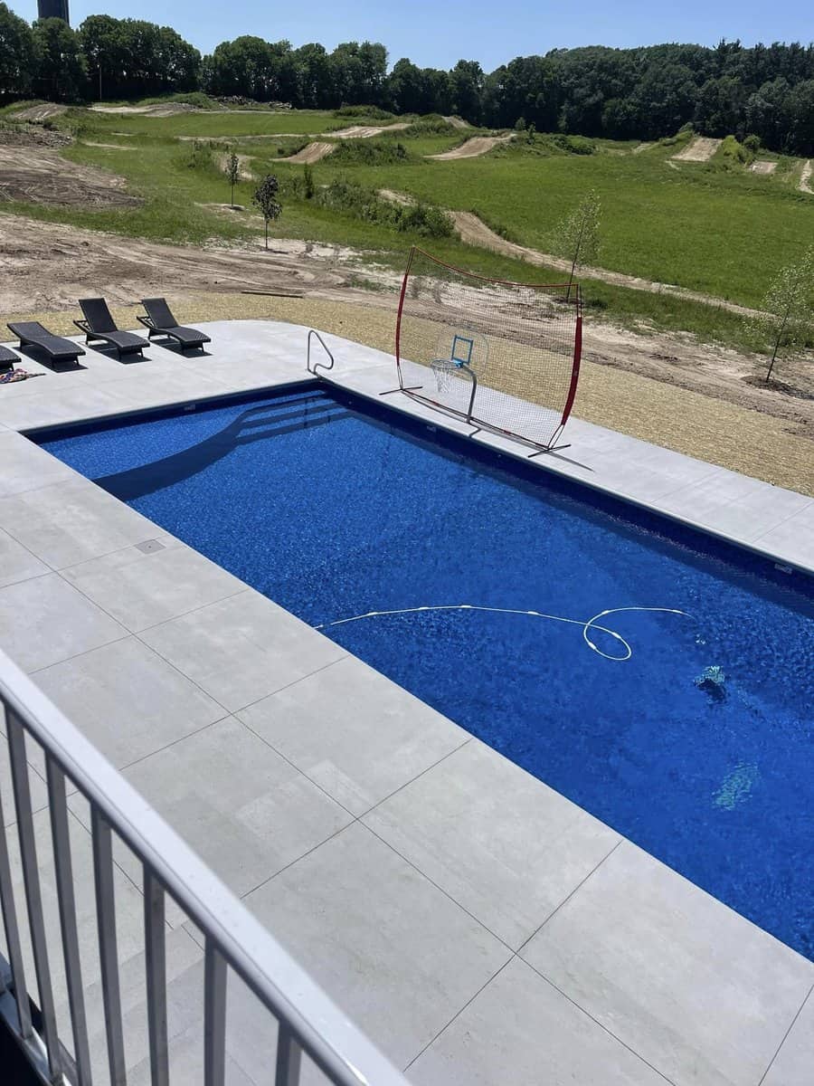 An outdoor swimming pool with blue water, surrounded by a concrete deck and loungers, with a natural landscape in the background.