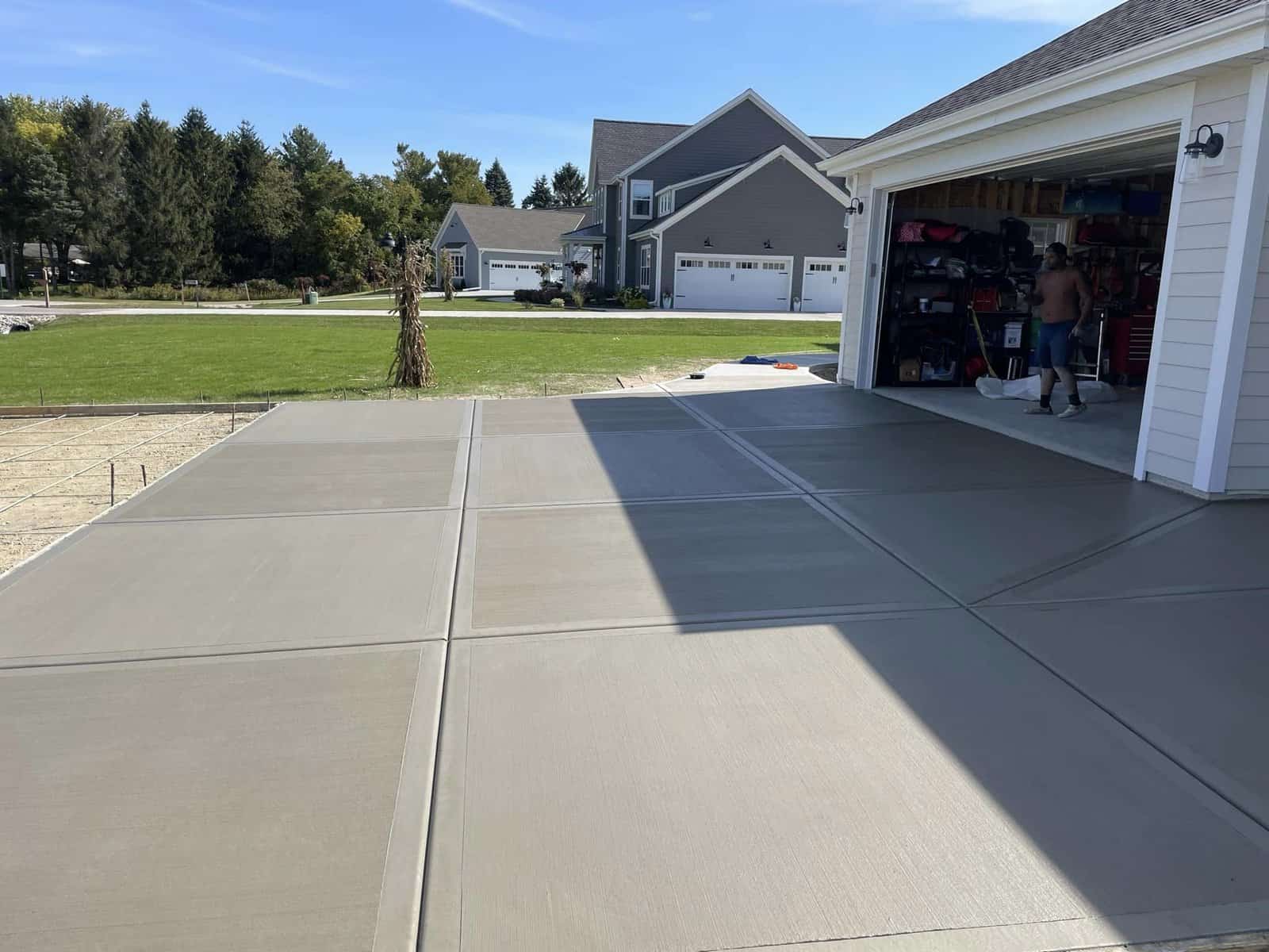 A fresh concrete driveway leading to an open garage with a person standing inside, set against the backdrop of a residential neighborhood with green lawns and houses under a clear blue sky.