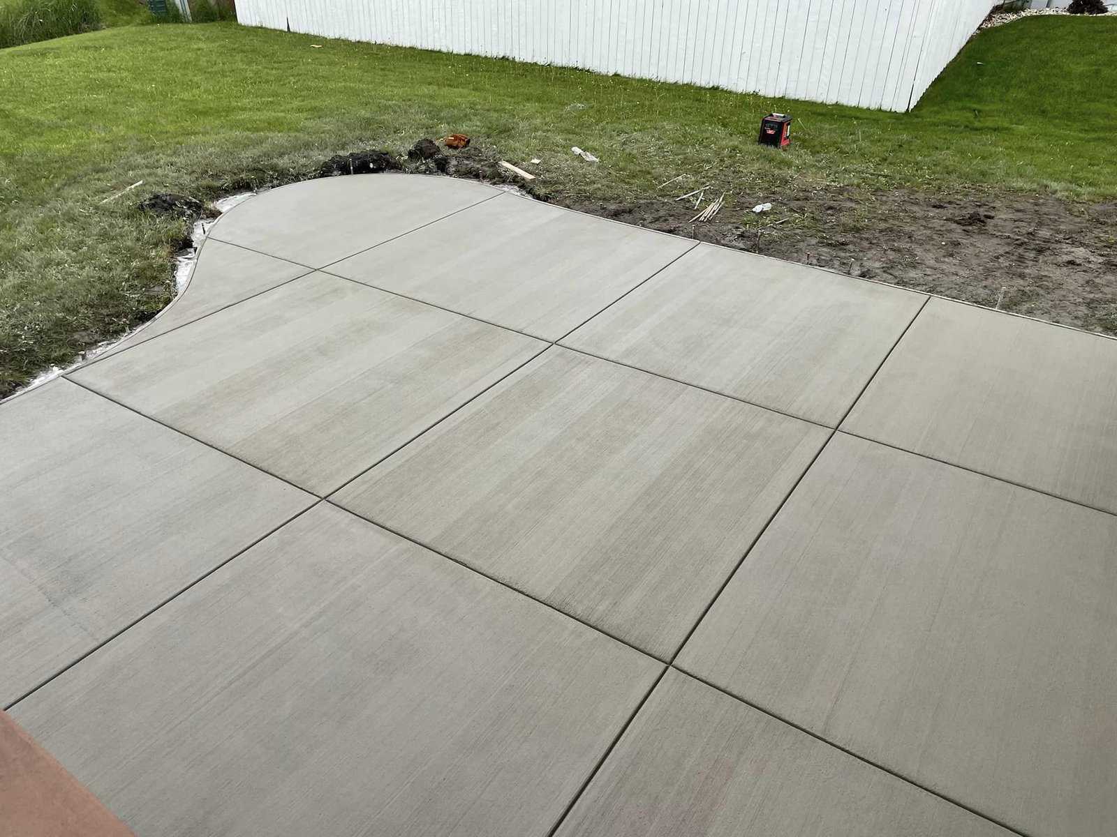 Newly poured concrete walkway with smooth finish and control joints, adjacent to a grassy area with gardening tools in the background.