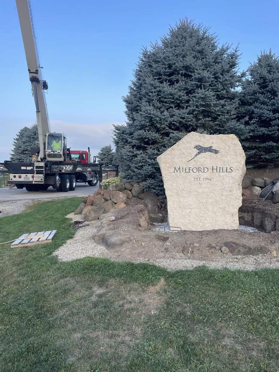 A crane truck parked beside a landscaped area with a large stone sign that reads "milford hills est. 1936".