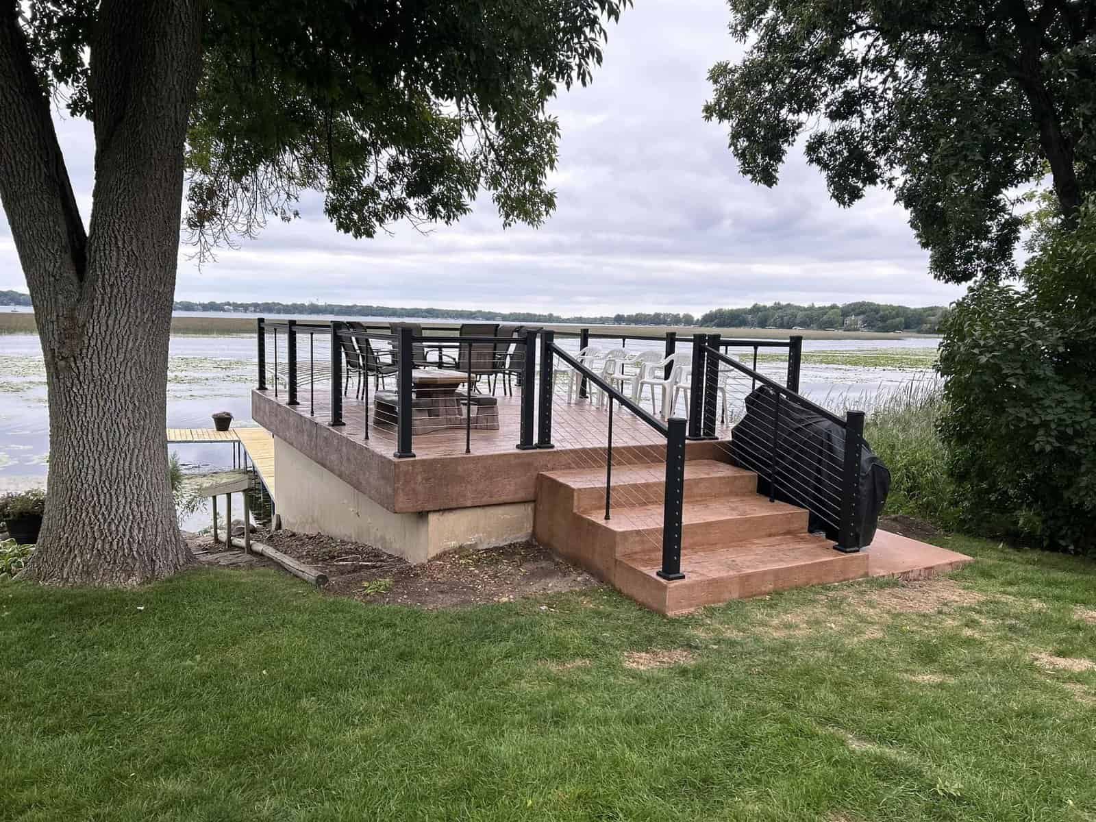 A lakeside wooden deck featuring a staircase, an assortment of chairs and a table, with a view of the water and overcast skies.