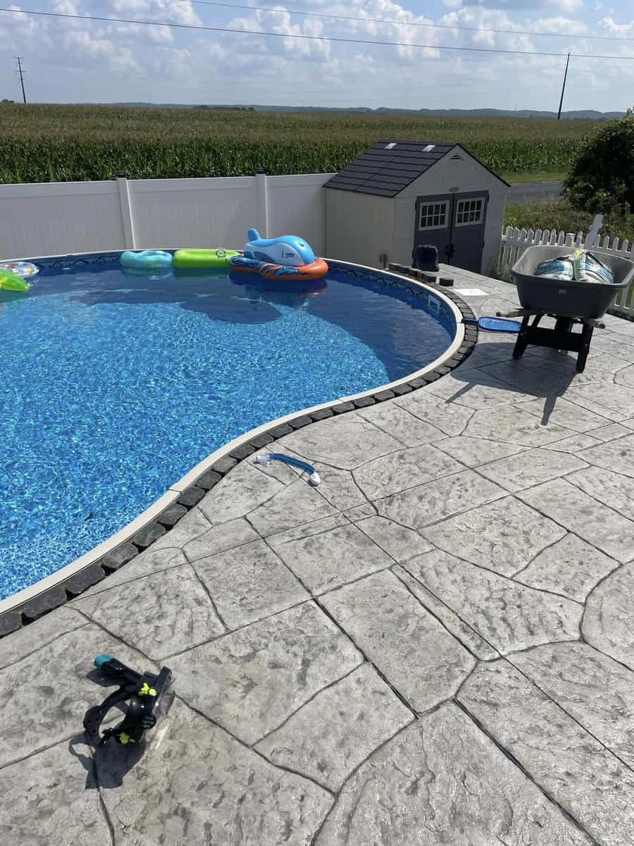 Above-ground swimming pool with inflatable toys, flanked by a small shed and a barbeque grill, on a patterned concrete patio.