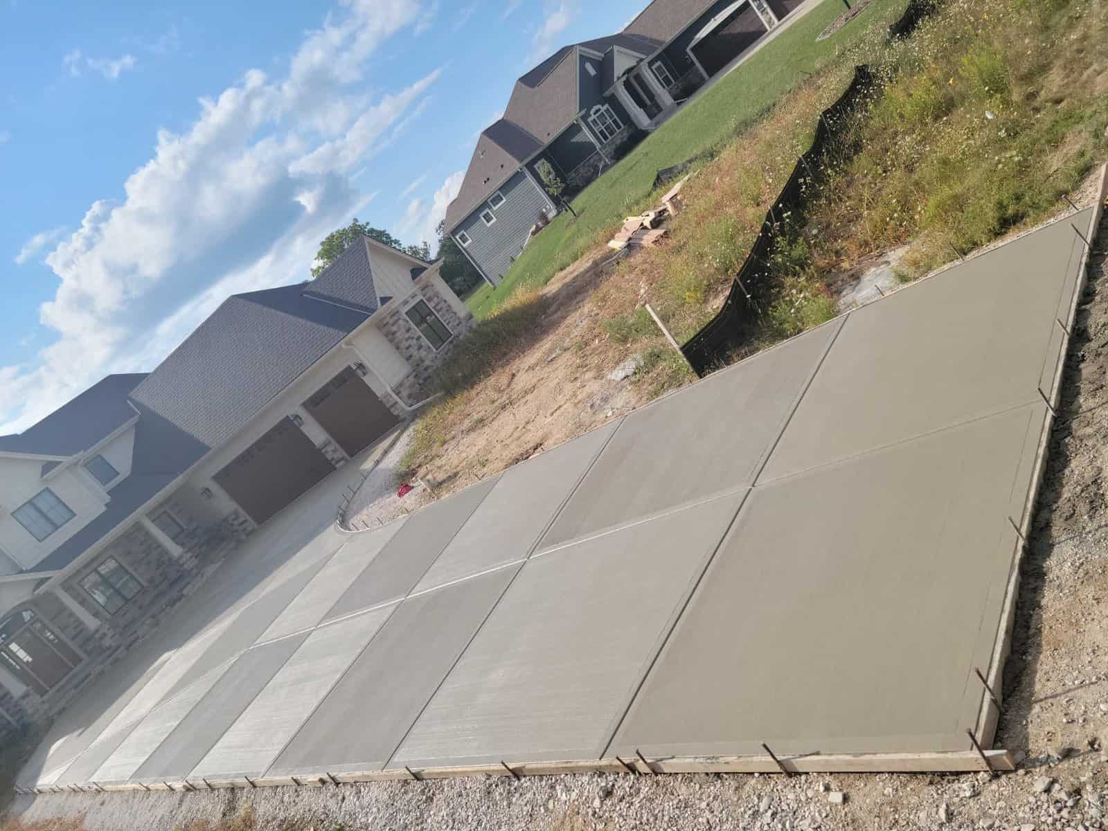 Newly poured residential concrete driveway with expansion joints, adjacent to a grassy area and in front of suburban homes under a clear sky.