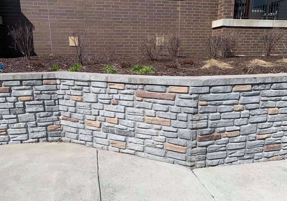 A stone retaining wall with mixed sizes of grey and beige bricks, alongside a concrete sidewalk and in front of a building with a brown brick facade.