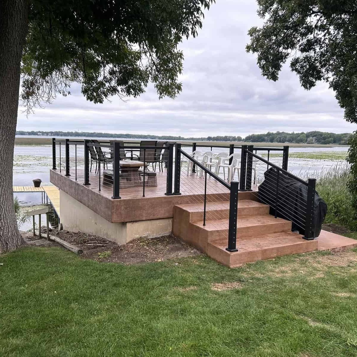 An outdoor patio with furniture overlooking a calm body of water, featuring steps leading down to a grassy area and a picturesque view of the treeline in the distance.